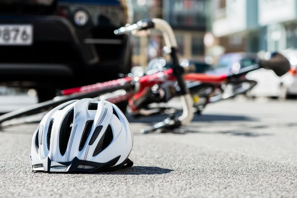 Close-up on white bike helmet lying on pavement with blurred red bike on its side in background abd black car visible behind it.